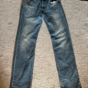 Levi's 501 blå jeans W32 L34 - Klassiska Levi's 501 jeans i blå denim med raka ben och normal passform. Jeansen har fem fickor, knappgylf och snyggt slitna detaljer. Tillverkade i bomull och märkta med Levi's Premium-label. Perfekta för en avslappnad och tidlös stil.