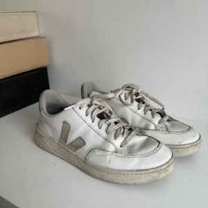 Vita sneakers från Veja - Säljer ett par klassiska vita sneakers från Veja med beige mockadetaljer och snörning. Skorna har en platt sula och rund tå, perfekt för en stilren vardagslook. Ikonisk V-logga på sidan och bekväm passform. Bra skick förutom sömnen som börjat släppa fram på skorna