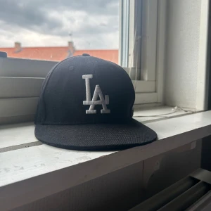 Svart LA-keps från New Era 59FIFTY - Säljer en svart keps från New Era 59FIFTY med LA-logga broderad framtill i vitt. Klassisk basebollmodell med platt skärm och MLB-logga baktill. Perfekt för dig som gillar sportig stil och streetwear.