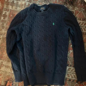 Mörkblå kabelstickad tröja Polo Ralph Lauren - Kabelstickad mörkblå tröja från Polo Ralph Lauren med klassisk rund halsringning och broderad grön logga på bröstet. Tröjan har långa ärmar och ribbade muddar för en snygg passform. Perfekt för dig som gillar stilren och tidlös design.