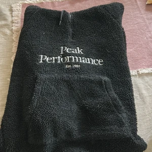 Svart fleece hoodie Peak Performance - Mysig svart fleece hoodie från Peak Performance med broderad logga på bröstet. Tröjan har en stor magficka, halv dragkedja och huva med snörning. Perfekt för kyliga dagar när du vill vara både bekväm och snygg.