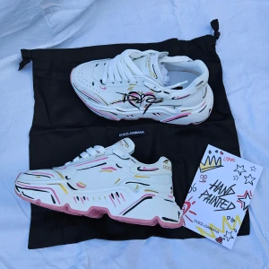 Dolce & Gabbana handmålade sneakers - Unika vita sneakers från Dolce & Gabbana med handmålat grafiskt mönster i svart, rosa och guld. Skorna har chunky rosa sula med logotyp, rund tå och snörning. Kommer med originalpåse och box.