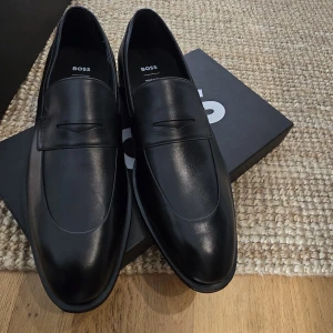Svarta loafers från BOSS - Säljer ett par stilrena svarta loafers från BOSS i skinn. Skorna har en klassisk design med rund tå och platt sula, perfekta för en dressad look. Diskret BOSS-logga på insidan och sula i läder. Passar perfekt inför bröllop studenten mm