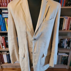 Beige linnékavaj med randigt foder - Inga defekter men använd! Stilren beige kavaj i linne med två knappar framtill och tre fickor. Insidan har ett vitt foder med tunna svarta ränder som ger en snygg kontrast. Nypris: 1900kr