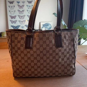 Gucci brun axelväska med GG-mönster - Snygg axelväska från Gucci i brunt GG-monogram. Väskan har två rejäla handtag i mörkbrunt skinn och detaljer i läder. Insidan har ett fack med dragkedja. Perfekt storlek för vardagens essentials och ger en lyxig touch till din outfit. Skriv för mer information🙌