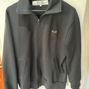 Svart jacka Comme des Garçons Play L - Svart jacka från Comme des Garçons Play med dragkedja, två fickor och ikoniskt hjärta med ögon på bröstet och stort tryck på ryggen. Jackan har hög krage och är i en loose passform, perfekt för en clean och stilren look. Hul i ärmen som också visas, skriv för mer information🙌