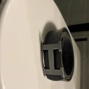 Svart skärp med H-spänne - Stilrent svart bälte i skinn med ett stort, silverfärgat H-format spänne. Perfekt accessoar för att lyfta din outfit med en clean och modern look. Bältet är brett och har en klassisk design som passar till jeans eller kostymbyxor.