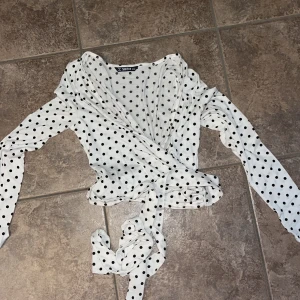 Prickig omlottopp från SHEIN XS - Superfin vit omlottopp från SHEIN med svarta prickar. Toppen har långa ärmar, djup v-ringning och knytband i midjan. Perfekt för dig som gillar en söt och trendig look. Tunn och mjuk i materialet, passar till många outfits.