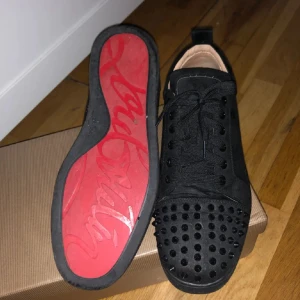Loubs svarta - Svarta sneakers från Christian Louboutin i mocka med coola svarta nitar på tån och klassisk röd sula. Skorna har snörning och rund tå, och insidan är fodrad i beige läder. Perfekta för dig som vill sticka ut med en edgy vibe. några spikar har ramlat av inget man tänker på 