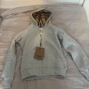 Grå hoodie från Burberry med klassiskt rutigt foder - Säljer en grå hoodie från Burberry med dragkedja och fickor. Insidan av huvan har det ikoniska Burberry-rutiga fodret i beige, rött och svart. Perfekt för en stilren och avslappnad look. Märkeslapp och originalpåse medföljer. PRIS KAN DISKUTERAS!!