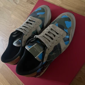 Valentino Rockrunner sneakers med camouflage - Säljer ett par Valentino Rockrunner sneakers med beige mockadetaljer och klassiskt camouflage-mönster i blått, grönt och svart. Skorna har snörning, rund tå och platt sula. Perfekta för dig som vill sticka ut med exklusiva sneakers.