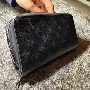 Svart Louis Vuitton plånbok i skinn - Säljer en svart och ganska stor plånbok från Louis Vuitton med klassiskt monogrammönster. Plånboken är tillverkad i skinn och har dragkedja runtom. Insidan har flera fack och detaljer i silverfärgad metall. Perfekt för dig som gillar lyxiga accessoarer. Skriv privat för mer information och bilder. Knappt använd och finns inga defekter.