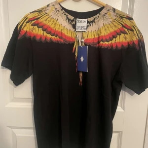 Svart t-shirt Marcelo Burlon med vingar - Svart t-shirt från Marcelo Burlon County of Milan med ikoniskt vingtryck i gult, rött och beige över axlar och hals. Klassisk passform och rund halsringning. Perfekt statement-plagg för dig som vill sticka ut.