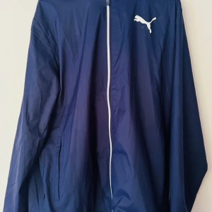 Blå vindjacka från Puma - Säljer en blå vindjacka från Puma med vit dragkedja och logga på bröstet samt ryggen. Jackan har huva och är tillverkad i lätt syntetmaterial, perfekt för blåsiga dagar. Klassisk sportig look med lång ärm och två fickor framtill.