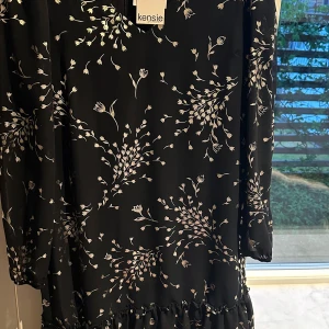 Svart blommig klänning från Kensie - Supersöt svart kortklänning från Kensie med vitt blommönster, v-ringning och volangkant nertill. Lätt och flowigt material, perfekt för dig som gillar en feminin och trendig stil. Klänningen har långa ärmar och en snygg passform som känns både chill och snygg.