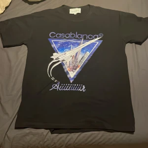 Svart Casablanca t-shirt med tryck - Svart t-shirt från Casablanca med stort futuristiskt tryck framtill som visar ett flygplan och stadssilhuett i blå och lila toner. Klassisk passform och rund hals. Perfekt statement-plagg för dig som gillar unika prints och streetwear-vibe.