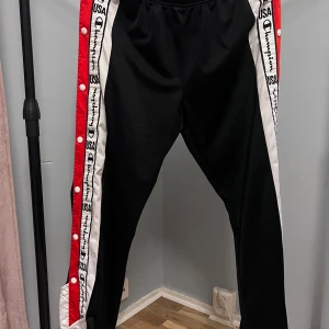 Champion svarta träningsbyxor L - Svarta träningsbyxor från Champion med röda och vita sidor, tryckknappar längs benen och logga på sidorna. Elastisk midja och sportig look, perfekta för chill eller träning. Materialet är polyester och byxorna har en loose passform.