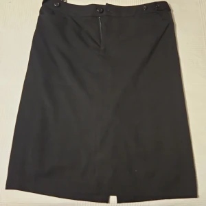 Svart pennkjol från Flash Classic - Klassisk svart pennkjol från Flash Classic i storlek 40. Kjolen har knappar i midjan och dragkedja bak för enkel påklädning. Perfekt för en stilren och enkel look.