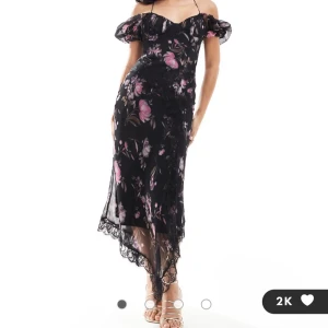 Svart blommig långklänning med spetsdetaljer - Romantisk svart långklänning med rosa och lila blommönster. Klänningen har offshoulder-ärmar, tunna axelband och spetsdetaljer längs nederkanten. Perfekt för dig som vill sticka ut med en feminin och elegant look. Helt ny med prislapp kvar. Säljer då den var för stor.