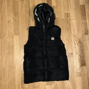 Svart dunväst med huva från Moncler - Snygg svart dunväst från Moncler med avtagbar huva och dragkedja framtill. Västen har två fickor med dragkedja och Moncler-logga på bröstet. Perfekt för lager-på-lager och streetstyle.