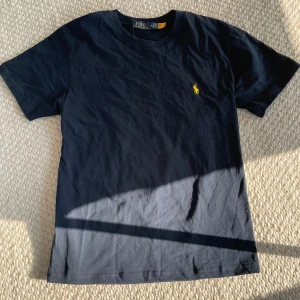 Marinblå t-shirt från Polo Ralph Lauren - Säljer en klassisk marinblå t-shirt från Polo Ralph Lauren med gul broderad logga på bröstet. T-shirten har rund halsringning och är tillverkad i mjuk bomull. Perfekt för en stilren och enkel look.