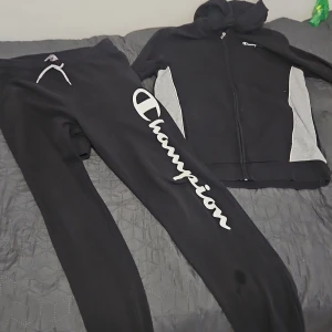 Svart hoodiejacka från Champion med grå detaljer - Svart hoodiejacka från Champion med dragkedja och grå paneler på sidorna. Jackan har huva och logga på bröstet. Perfekt för en sportig och avslappnad stil.