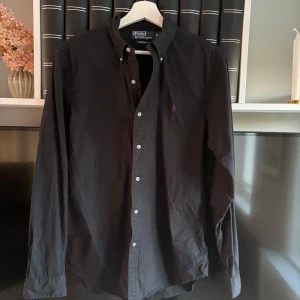 Svart skjorta från Ralph Lauren - Klassisk svart skjorta från Ralph Lauren med lila broderad logga på bröstet. Skjortan har knappar framtill och på kragen samt långa ärmar med manschettknappar. Perfekt för en stilren look. Skriv för frågor och funderingar !!!