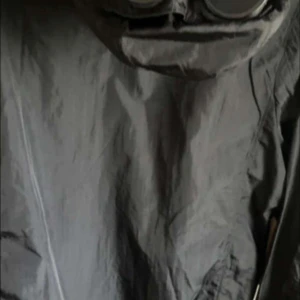 Svart vindjacka med goggles från C.P. Company - Säljer en svart vindjacka från C.P. Company med ikoniska goggles i huvan och dragkedja framtill. Jackan har en sportig look och är perfekt för dig som gillar streetwear. Justerbar huva och diskreta bröstfickor. Perfekt till fotbollsmatcher!