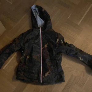 Camouflage vindjacka med huva från Moncler - Säljer en snygg vindjacka från Moncler med camouflagemönster och huva. Jackan har dragkedja framtill och är perfekt för dig som vill ha en cool och funktionell look. Moncler-logga på ärmen ger en exklusiv touch.
