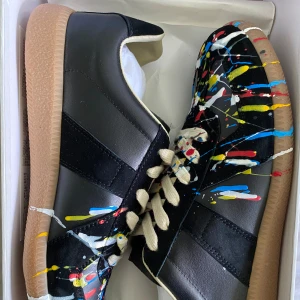 Maison Margiela med paint splatter - Unika svarta sneakers med färgstänk i blått, gult, vitt och rött. Skorna är i nytt skick och säljs då de är för små! Perfekt för dig som vill sticka ut med en kreativ och konstnärlig stil. Hör av dig vid minsta fråga!