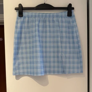 Blå rutig kortkjol - 🌸Säljer en söt blå kortkjol med vitt rutmönster. Rak passform och resår i midjan. Storleken är L men jag skulle säga att den snarare är S/M 🌸
