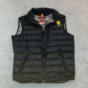 Svart dunväst från Parajumpers - Snygg svart dunväst från Parajumpers med dragkedja framtill och två fickor med dragkedja. Västen har en hög krage och klassisk gul detalj vid halsen. Perfekt för lager-på-lager och håller dig varm under kyliga dagar. ÄKTA! Nypris 4000