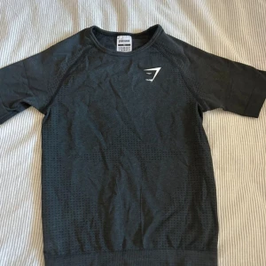 t-shirt från Gymshark !compression¡ - Säljer en mörkgrå t-shirt från Gymshark med diskret mönster och logga på bröstet. T-shirten har korta ärmar och är tillverkad i ett mjukt, stretchigt material som passar perfekt till träning. T-shirten är compression och visar fina konturer på kroppen🩷