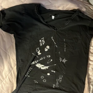 Svart t-shirt med tryck och v-ringning - Svart t-shirt i medium med coolt vitt tryck framtill, v-ringad hals och bröstficka. T-shirten har korta ärmar och en avslappnad passform. Perfekt för dig som gillar en edgy och unik stil.