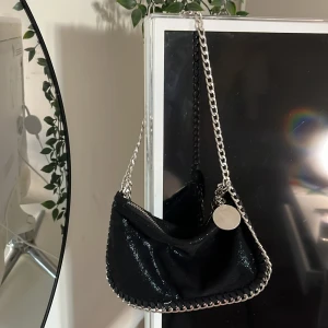 Stella McCartney handväska  - Snygg svart axelväska med glansig finish och silverfärgad kedja som axelrem. Väskan har en rundad form, dragkedja upptill och dekorativ kedja längs kanterna. Perfekt för dig som gillar en edgy och trendig look.