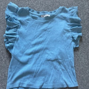 Blå ribbad topp med volangärmar från Zara - Supersöt blå ribbad topp från Zara med korta volangärmar. Toppen har rund halsringning och är tillverkad i ett mjukt, stretchigt material. Perfekt för dig som gillar en feminin och trendig stil.
