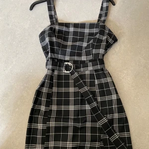 Svartvit rutig klänning med bälte från H&M Divided - Säljer en svartvit rutig klänning från H&M Divided med breda axelband och tillhörande bälte med silverfärgat spänne. Klänningen har en rak passform och är perfekt för dig som gillar stilrena mönster.