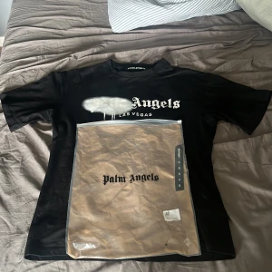 Svart Palm Angels t-shirt Las Vegas - Svart t-shirt från Palm Angels med tryck 'Las Vegas' och vit spray-effekt över loggan. Klassisk passform, rund hals och korta ärmar. Tillverkad i mjuk bomull och levereras med originalpåse. Perfekt för dig som gillar streetwear och statement-plagg.