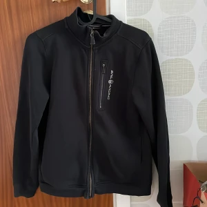 Svart jacka Sail Racing strl 160 - Svart jacka från Sail Racing med hel dragkedja och en bröstficka med logga och dragkedja. Tröjan har hög krage och är tillverkad i ett mjukt, syntetiskt material. Har en liten skada men är igensyd inget som syns när tröjan sitter på