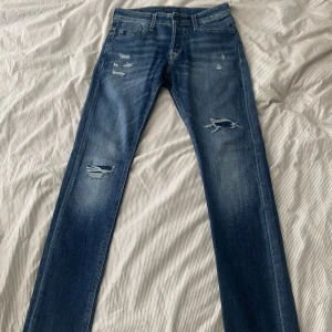 Slim Glenn blå slitna jeans Jack & Jones - Stilrena  jeans från Jack & Jones, modell Slim Glenn. Jeansen har slitna detaljer och hål på benen för en cool look. De är slim fit med klassisk femficksdesign och tillverkade i stretchig bomull för extra komfort.