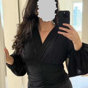 Svart smockad topp  - Säljer en svart smockad topp med djup v-ringning och följsamma ärmar. Toppen har snygg rynkdetalj framtill och markerad midja som ger en smickrande siluett. Inuti toppen finns inbyggt stöd för bysten. Perfekt att styla med jeans eller kjol, denna topp är otroligt smickrande. Storlek 38, endast använd en gång.