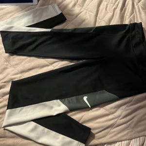 Nike leggings med svart och vitt på sidan - Snygga leggings från Nike i svart med grå och vita detaljer längs benen. De sitter mid-waist och är tillverkade i ett stretchigt syntetmaterial som passar perfekt för träning. Nike-loggan syns tydligt på ena benet. Riktigt sportig vibe!