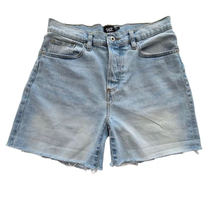 Ljusblå jeansshorts - Ljusblå jeansshorts med klassisk femficksdesign, råa kanter och normal passform. Shortsen har bälteshällor och knappgylf, perfekt för en chill sommarlook. Materialet är mjukt denim som ger en avslappnad vibe.