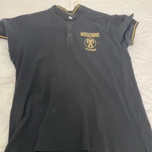 Svart pikétröja Moschino Swim XL - Svart pikétröja från Moschino Swim med gul logga och tryck på bröstet. Tröjan har korta ärmar, knappar vid halsen och gul detalj på krage och ärmslut. Perfekt för dig som gillar street och vill sticka ut med en snygg designerpiké.