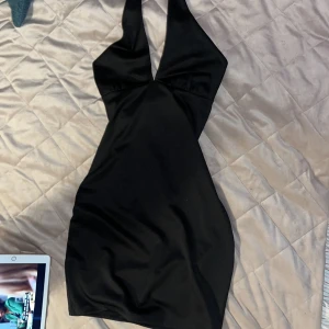 Svart halterneckklänning H&M Divided - Snygg svart kortklänning från H&M Divided med halterneck och djup v-ringning. Klänningen har en tight passform och är tillverkad i ett glansigt, stretchigt material som ger en stilren look. Perfekt för dig som vill sticka ut med en enkel men trendig stil.