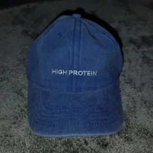 Blå keps Star Nutrition HIGH PROTEIN - Snygg blå keps från Star Nutrition med broderad text 'HIGH PROTEIN' framtill och 'WHEY-80' på sidan. Justerbar rem baktill med metallspänne. Tillverkad i bomull, perfekt för dig som gillar gym och träning.