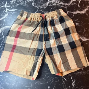 Beige rutig shorts från Burberry - Snygga shorts från Burberry i klassiskt beige rutigt mönster med svarta, röda och vita detaljer. Shortsen har elastisk midja med snörning och är tillverkade i ett lätt och bekvämt material. Perfekta för sommardagar när du vill sticka ut lite extra.