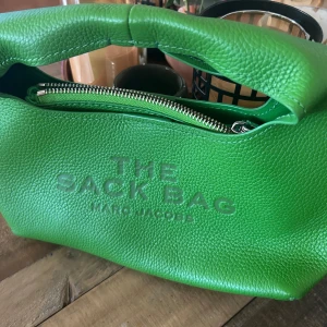 Marc Jacobs The Mini Sac Bag -Kiwi - Cool grön handväska 'The Mini Sack Bag' från Marc Jacobs i skinn med präglad logga och dragkedja. Tillhörande grön kuvertväska i textil och skinn med handledsrem och dragkedja. Perfekt för dig som vill sticka ut med färg!Viktiga egenskaper: Kiwi | Grön | 100% Koskinn | Magnetisk snäppstängning | Plånbok med dragkedja för säker förvaring | Minimalistisk design | Mått och passform: 24 cm L x 13 cm H x 14 cm W | Handtagshöjd: 12 cm | Material och skötsel: 100% Koskinn. Kvitto finns ej kvar.