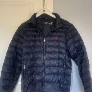 pufferjacka från Polo Ralph Lauren - Säljer en mörkblå pufferjacka från Polo Ralph Lauren med röd broderad logga på bröstet. Jackan har dragkedja framtill och två fickor med dragkedja. Perfekt för kyligare dagar, pris kan diskuteras!