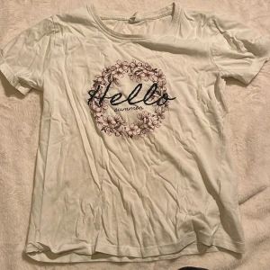 Vit t-shirt med blommor från Uniq Young - Vit t-shirt från Uniq Young med tryck av blommor i en cirkel och texten 'Hello summer' på bröstet. Klassisk rund halsringning och korta ärmar. Perfekt för dig som gillar en söt och somrig stil.